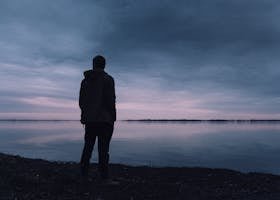 A solitary silhouette of a man in a jacket gazing at a lake during a peaceful sunset, creating a serene atmosphere.